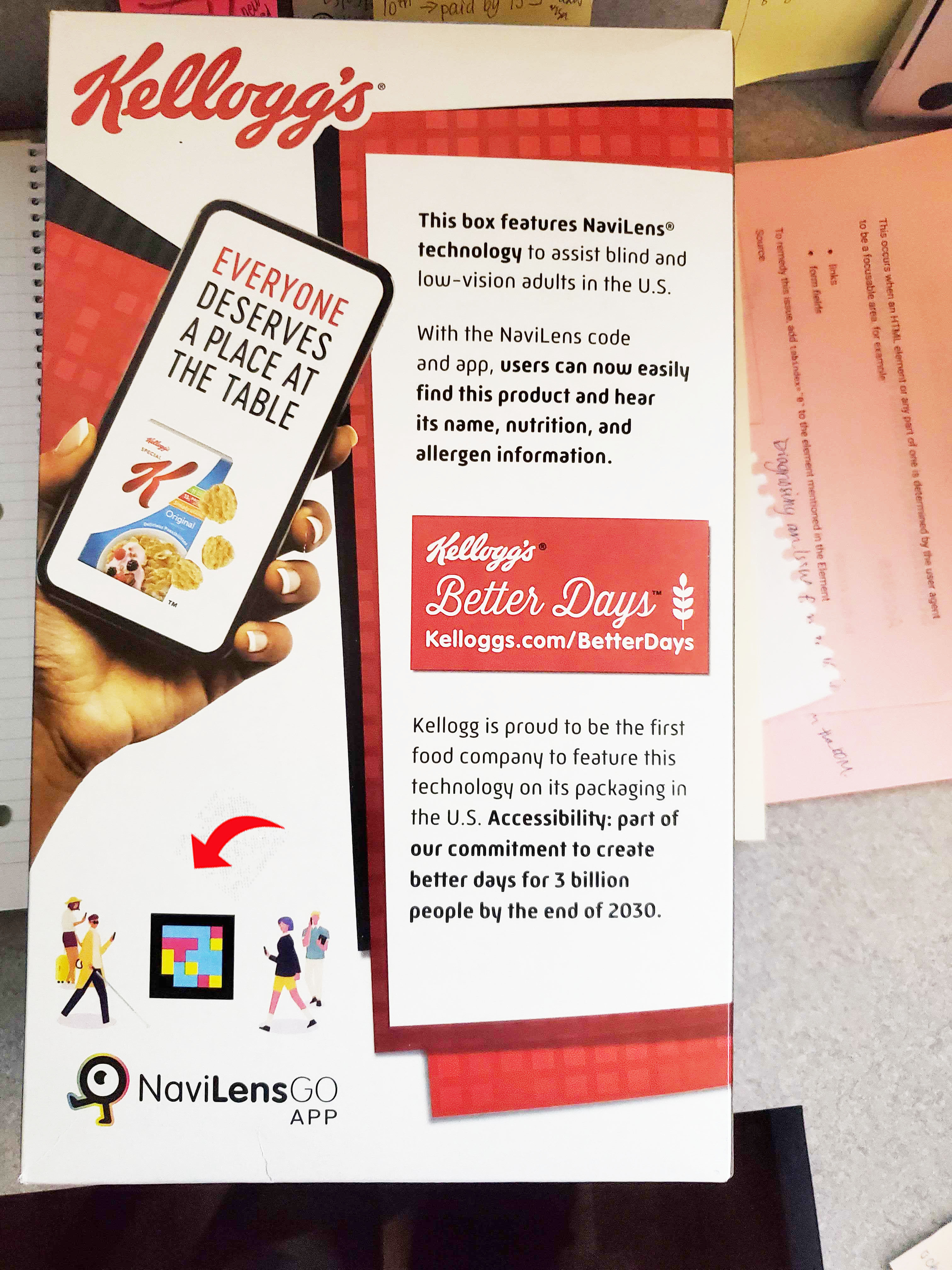 A Kellogg's cereal box with the Kellogg logo at the top. Below the logo is a hand holding a cell phone. On the cell phone screen a sentence reads - Everyone Deserves a Place at the Table. Next to the cell phone is a white panel with black text. The text reads - This box features NaviLens technology to assist blind and low-vision adults in the U.S. with NaviLens code and app, user can now easily find this product and hear its name, nutrition and allergen information. Kellog is proud to be the first food company to feature this technology on its packaging in the U.S. Accessibility: part of our commitment to create better days for 3 billion people by the end of 2030.